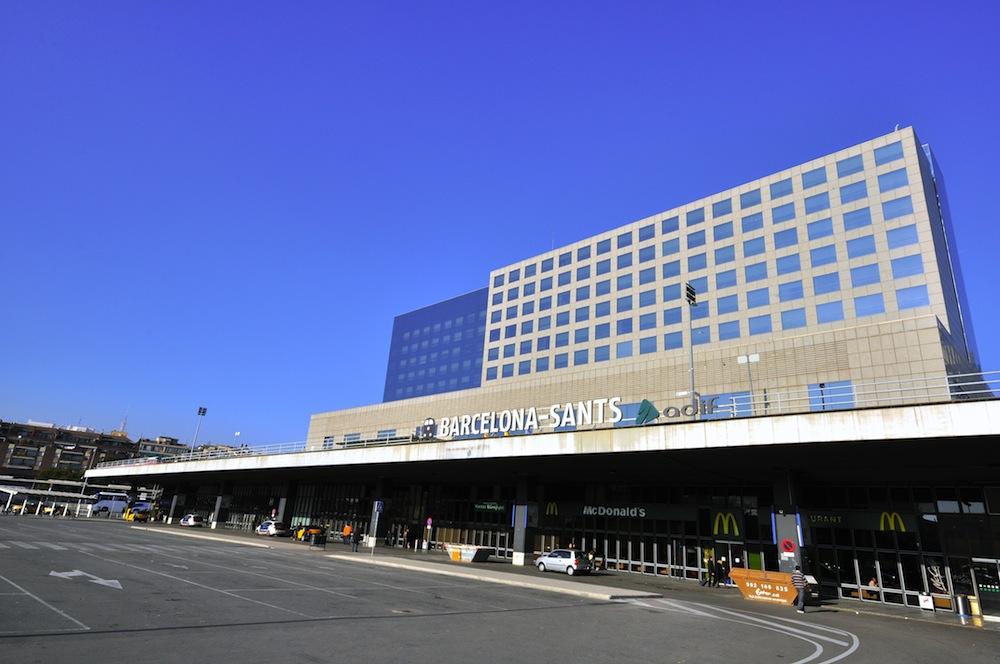 Fachada del estadio Barcelona Sants con letrero, estacionamiento y edificios bajo cielo azul despejado.
