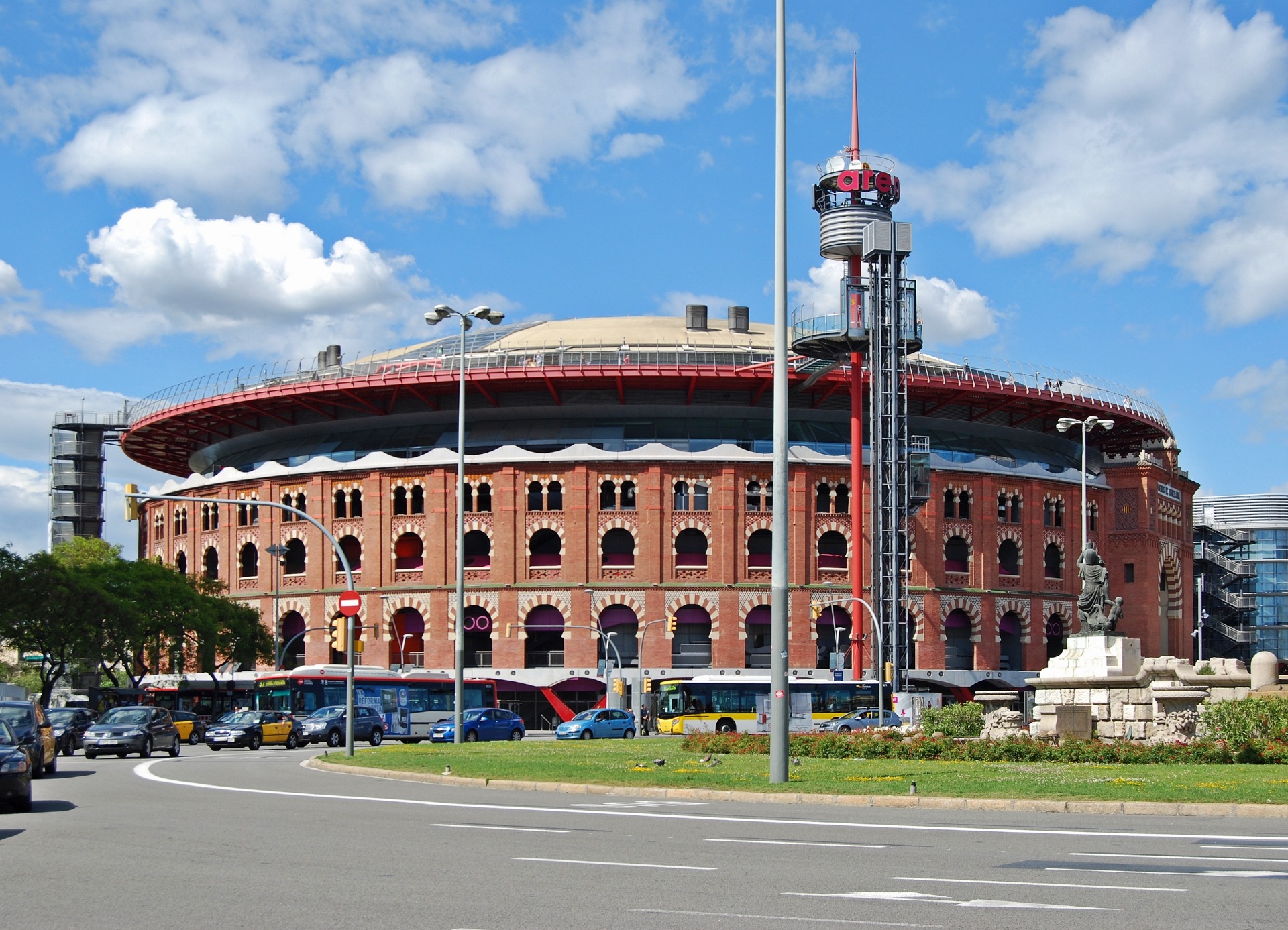 Estadio de ladrillo con techo rojo, torre de iluminación y tráfico de autobuses bajo cielo azul con nubes.
